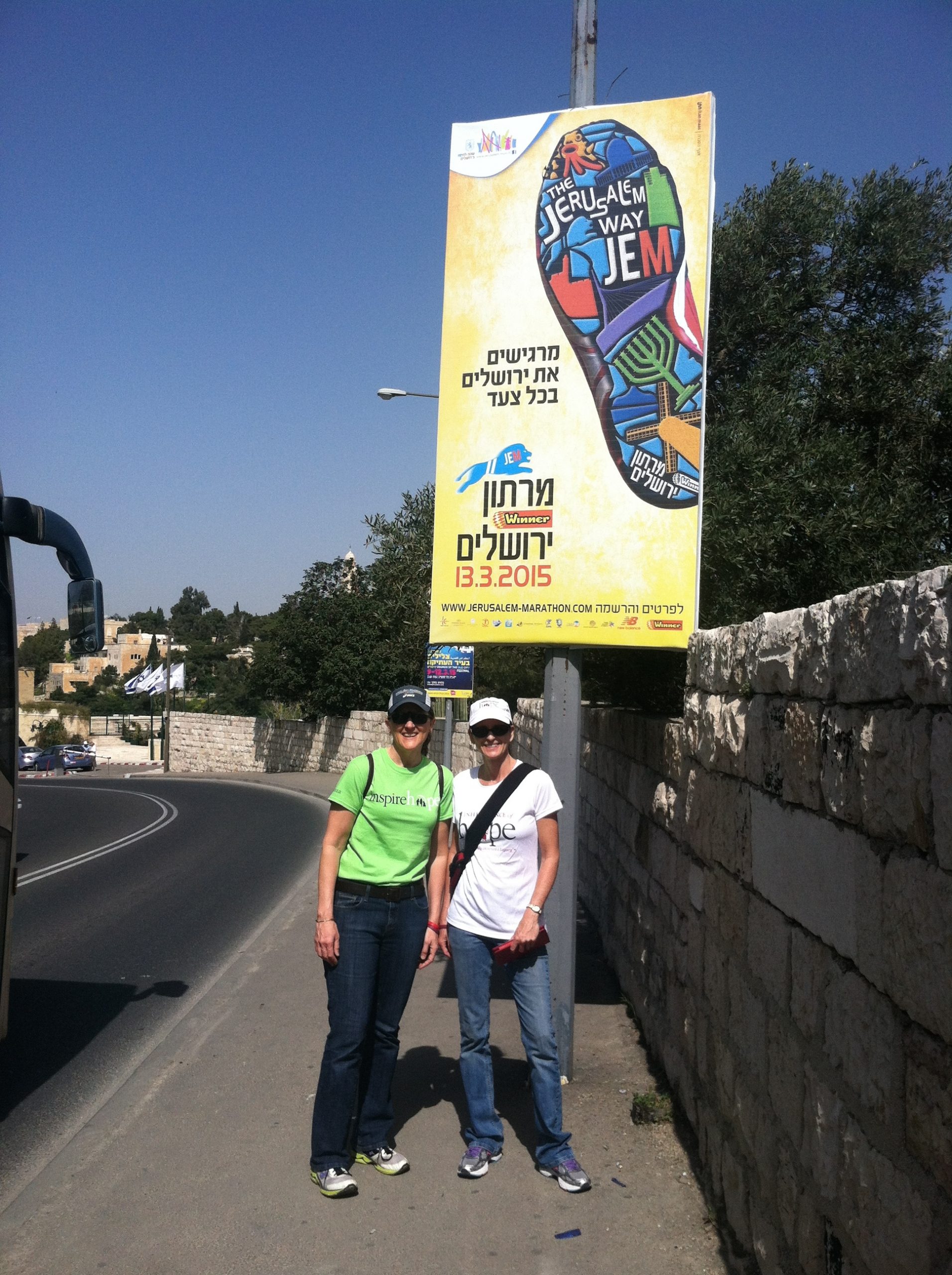 Two women smiling while standing in front of a sign with foreign text on the side of a winding road