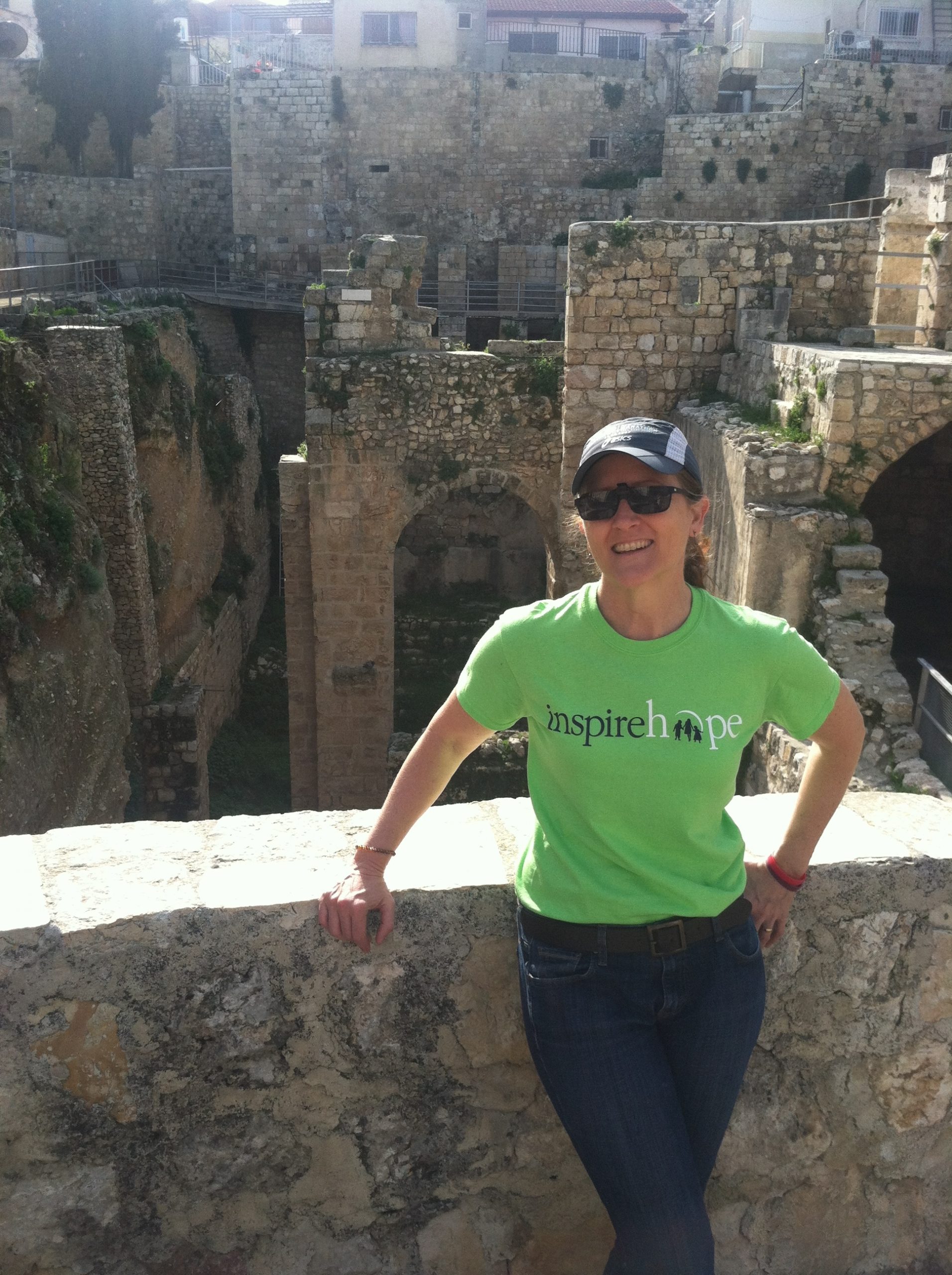 A woman wearing a green Inheritance of Hope t-shirt smiling while standing by a ledge with several archaic buildings in the background