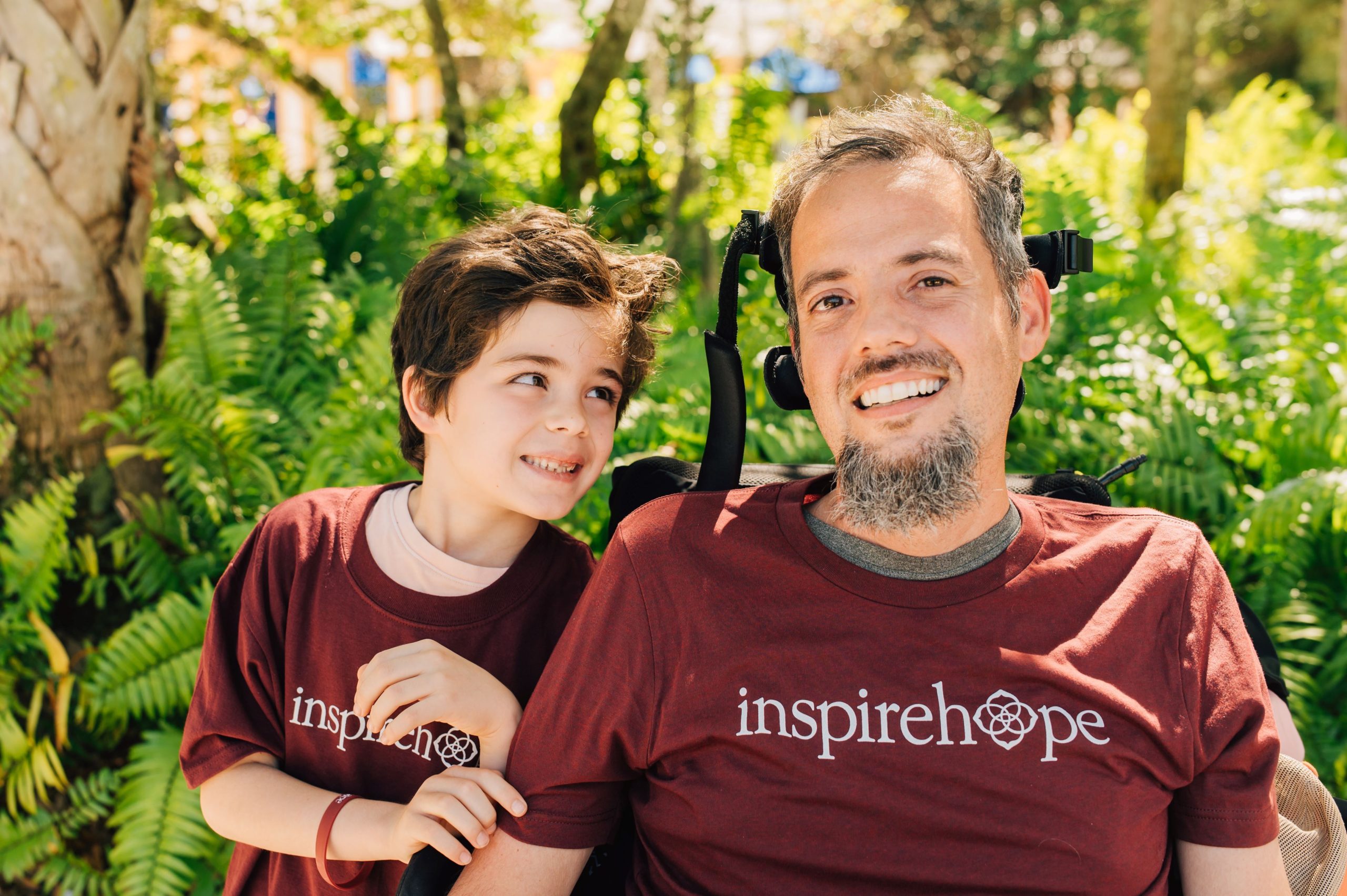 A wheelchair-bound man and young boy smiling with vibrant foliage in the background