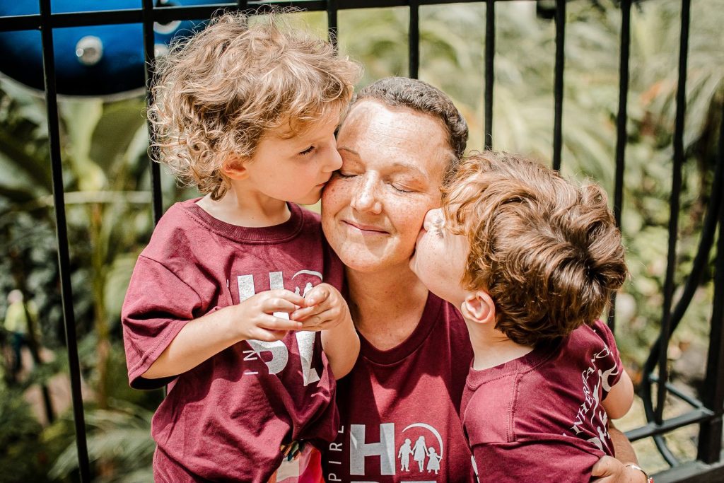 A mother wearing an Inheritance of Hope shirt closes her eyes and smiles as her two young children, also in matching shirts, give her kisses on the cheeks.