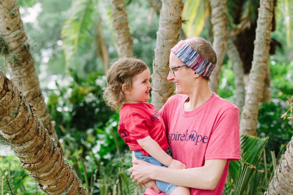 A mother and her young daughter, both wearing matching pink "Inspire Hope" shirts, share a joyful moment outdoors surrounded by greenery and palm trees.