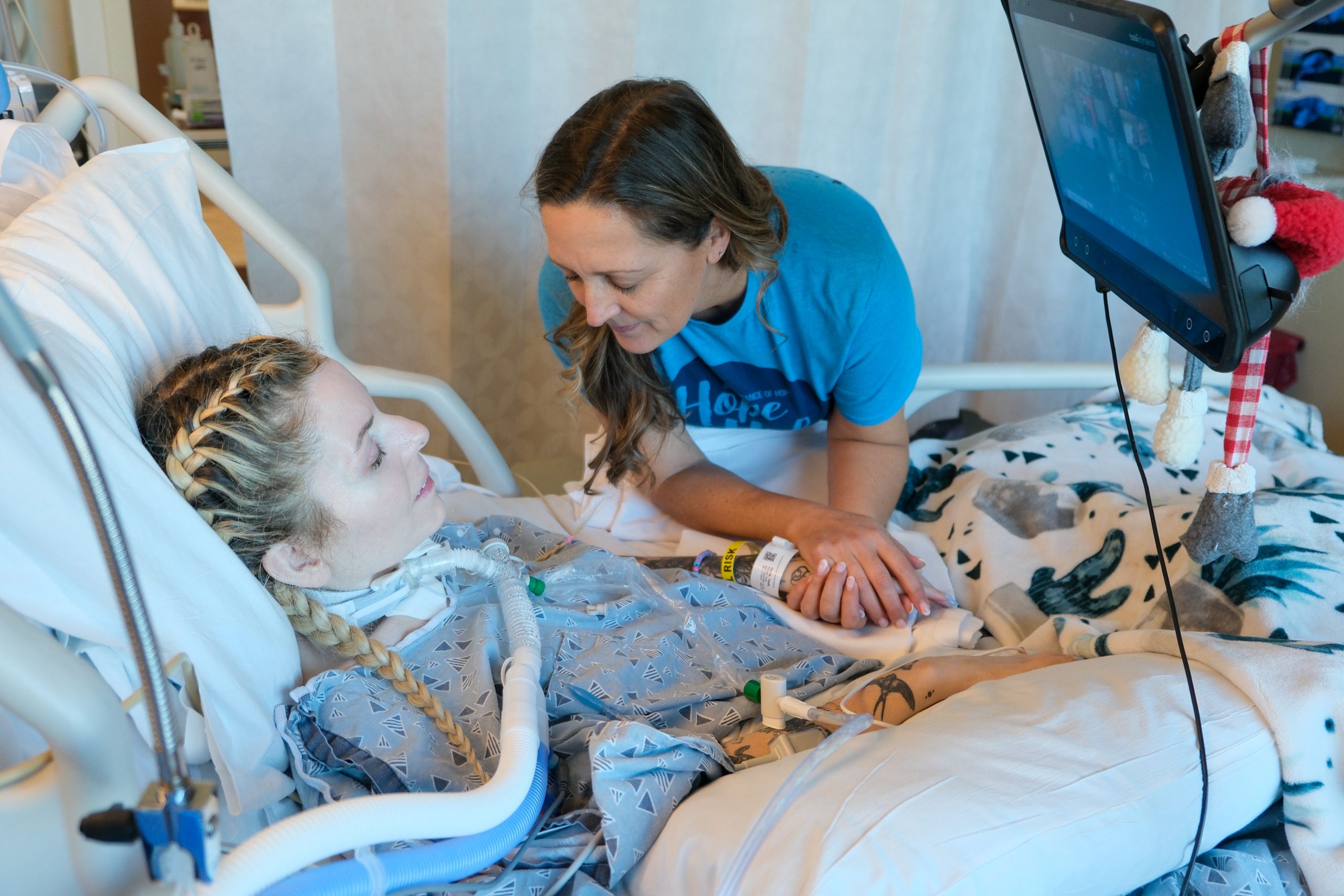 A woman in a blue Inheritance of Hope shirt holds hands and prays with a Hope Hub participant in a hospital bed.