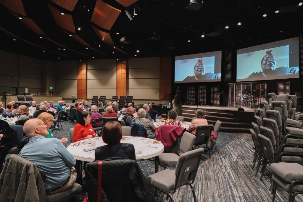 A group of senior adults sit at round tables in a church auditorium watching a testimonial about Legacy Videos