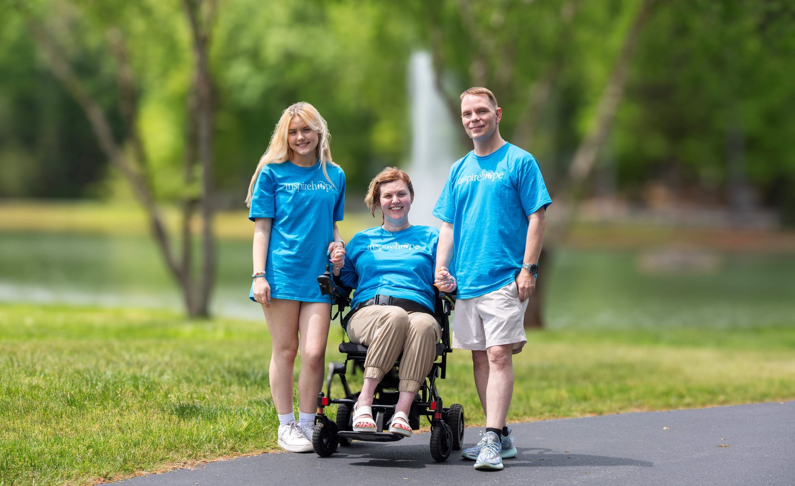 Photo of a young mom with ALS in an electric wheelchair. Beside her is her husband and daughter holding her hands outside.