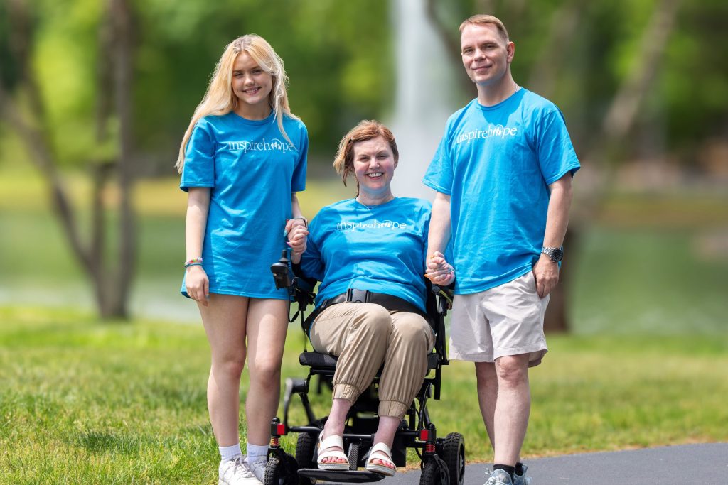 Photo of a young mom with ALS in an electric wheelchair. Beside her is her husband and daughter holding her hands outside.