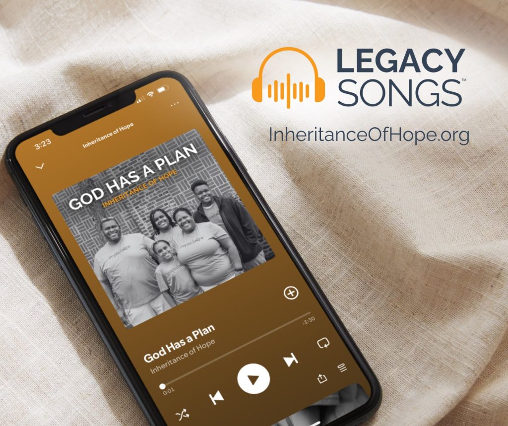 A phone resting on a light cloth shows a song titled “God Has a Plan” by Inheritance of Hope, with a black-and-white photo of a smiling family. Next to the phone is the Legacy Songs logo and the website InheritanceOfHope.org.
