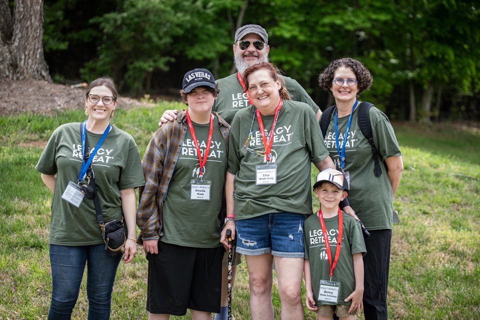 A smiling family and two Inheritance of Hope volunteers wearing matching "Legacy Retreat" shirts pose outdoors in front of a lake and fountain.