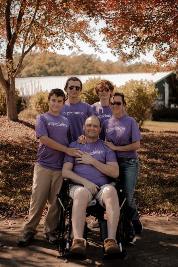 A young father in a wheelchair is embraced by his family. Surrounding them is fall foliage.