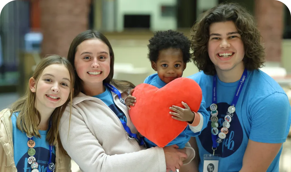 A toddler holding a stuffed heart being embraced and surrounded by volunteers