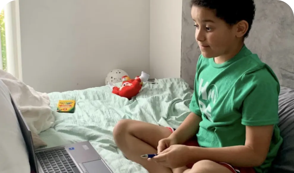 A young boy sitting on a bed looking at an open laptop