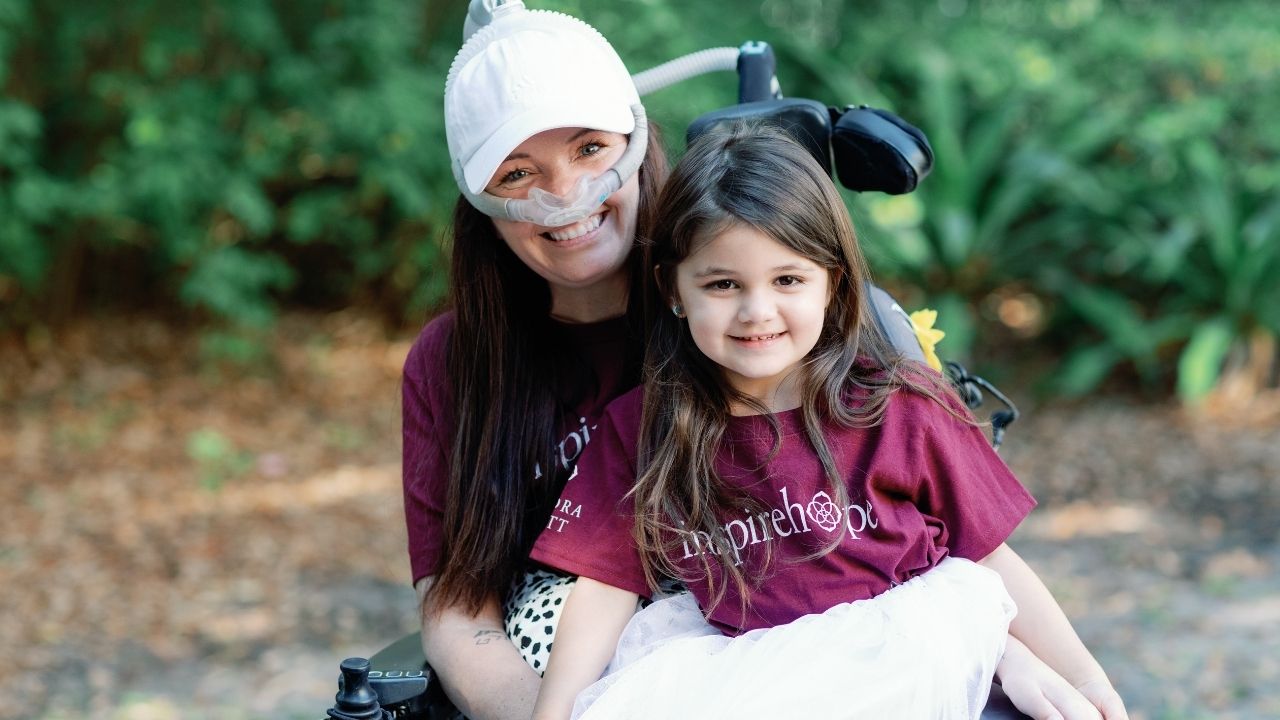 A woman in a motorized wheelchair wearing a nose respirator smiling with a young girl sitting on her lap