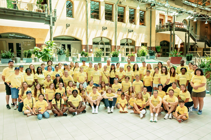 “New memories, lifelong bonds.” A large group of Inheritance of Hope members wearing yellow t-shirts smiling for a picture inside a facility