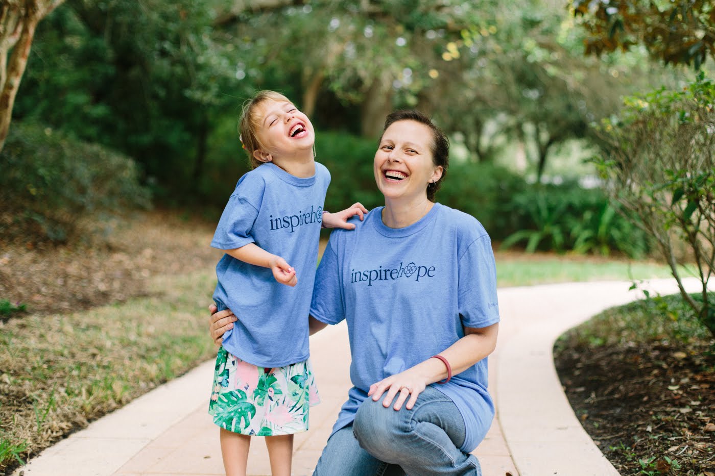 A mother smiling and kneeling next to her daughter on a walkway with foliage in the background