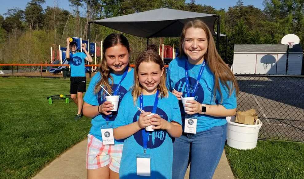 A young woman and two young girls standing shoulder to shoulder smiling with a playground in the background