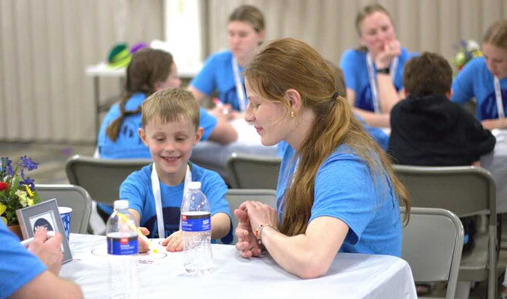 A young boy coloring at a table with a young woman supervising him. Several others are doing arts and crafts in the background