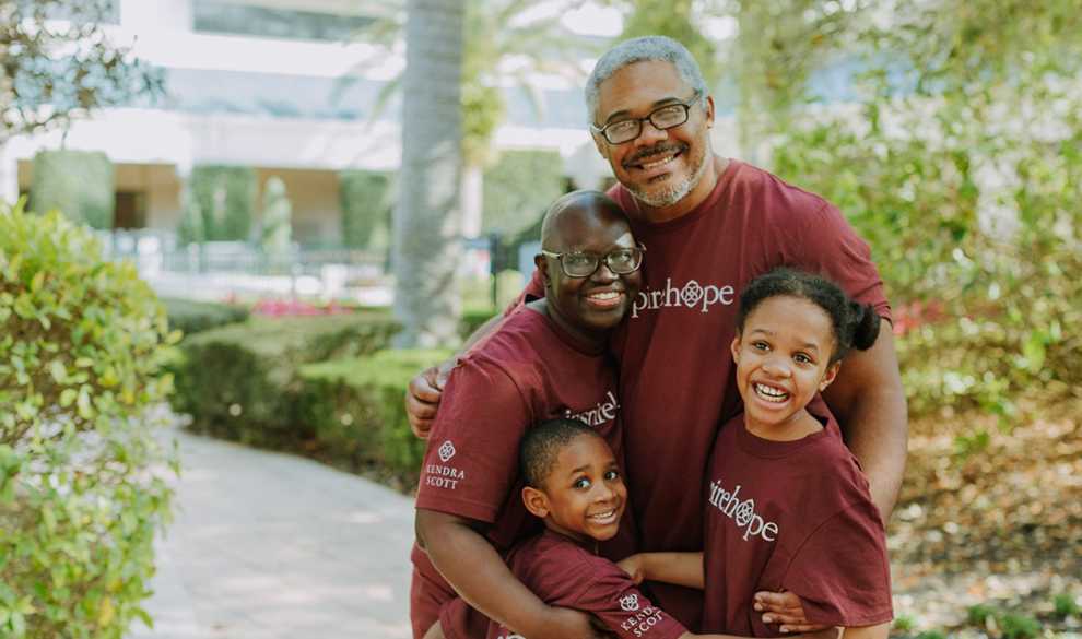A mother and father smiling and embracing their two children. A walkway and foliage are in the background