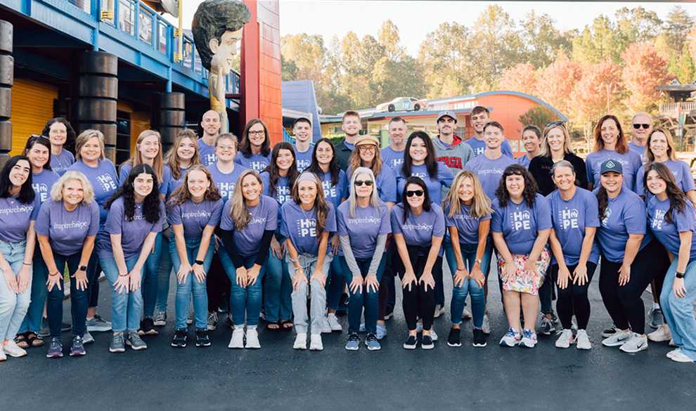 supportive_community A large group of Inheritance of Hope members wearing purple t-shirts smiling for a picture at an amusement park