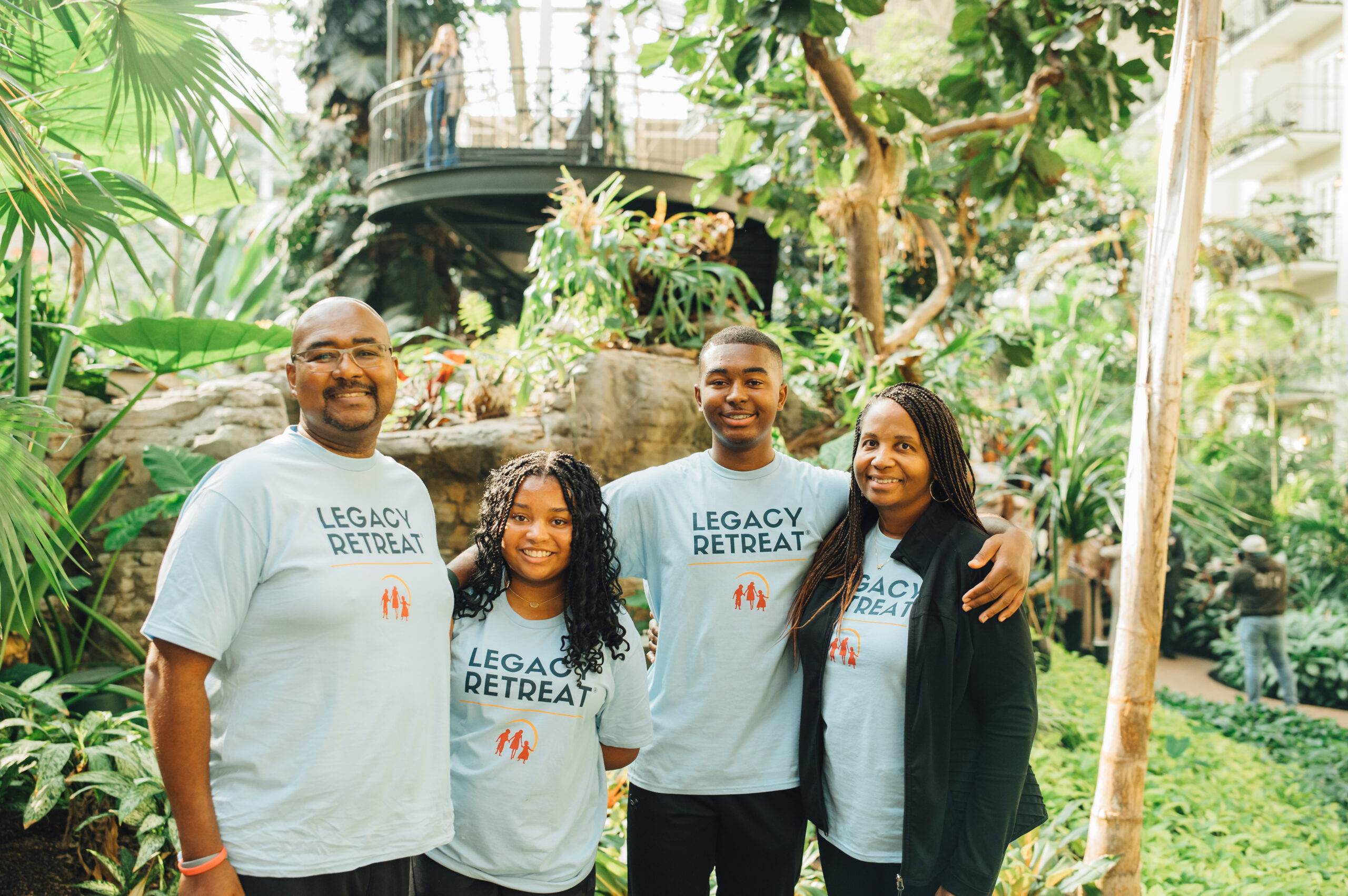 A family wearing matching Legacy Retreat shirts smiles together in a lush indoor garden.
