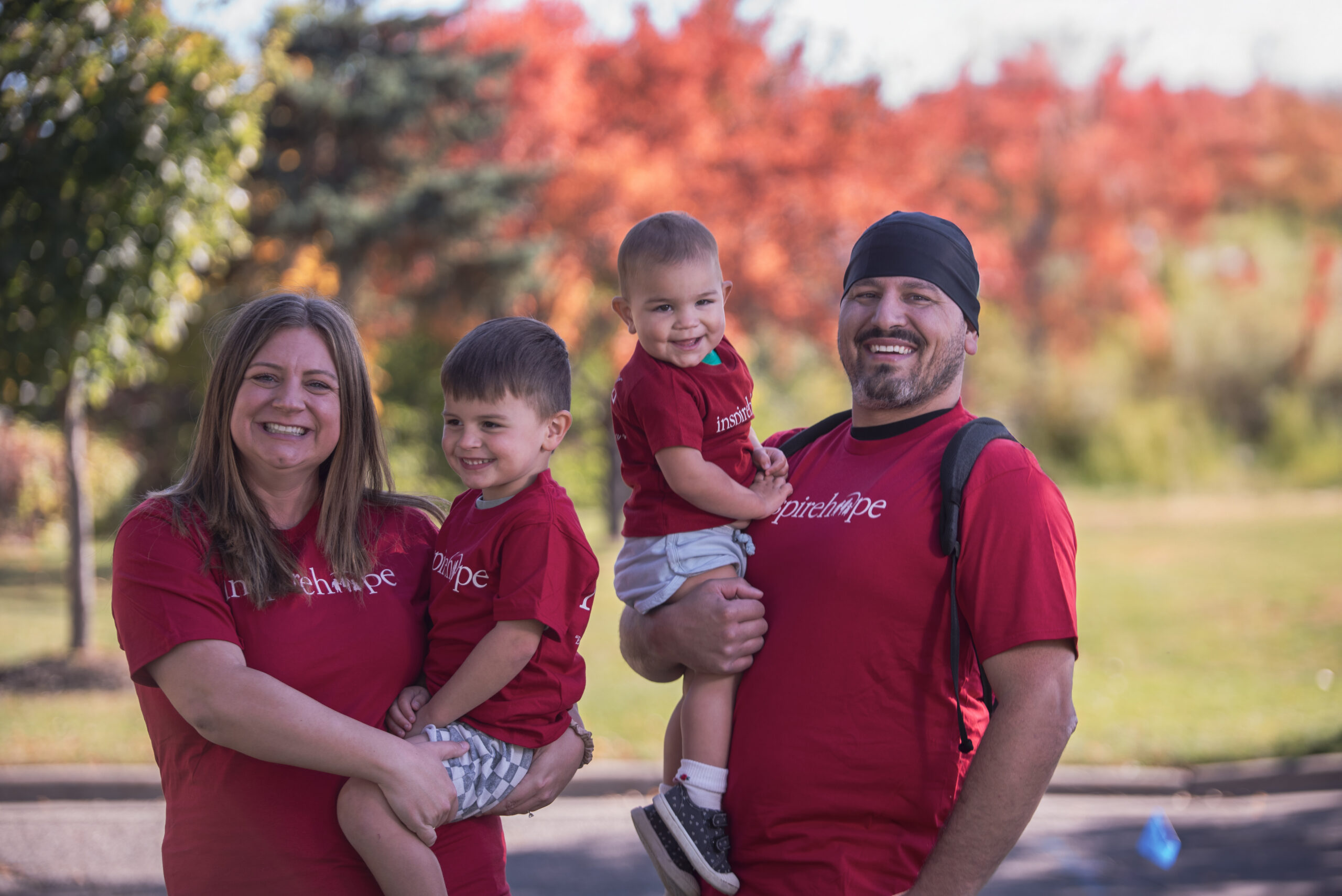 A smiling family of four wearing matching red shirts poses outdoors together with colorful autumn trees in the background.